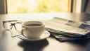 A table with a cup of coffee, pile of newspapers and set of glasses, with sunlight coming in from window behind